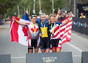 Image of Soldier On Ambassador Sarah Watson wearing a gold Invictus Games Medal standing between two other Medal winning competitors. Sara is wearing an Invictus Australia competitors uniform and is holding and Australian flag behind her back