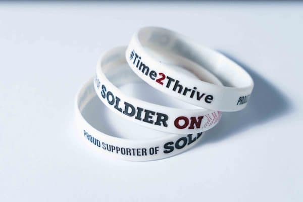 Three stacked white wristbands with text 'Time 2 Thrive', and 'Proud Supporter of Soldier On' in dark grey and red on them.